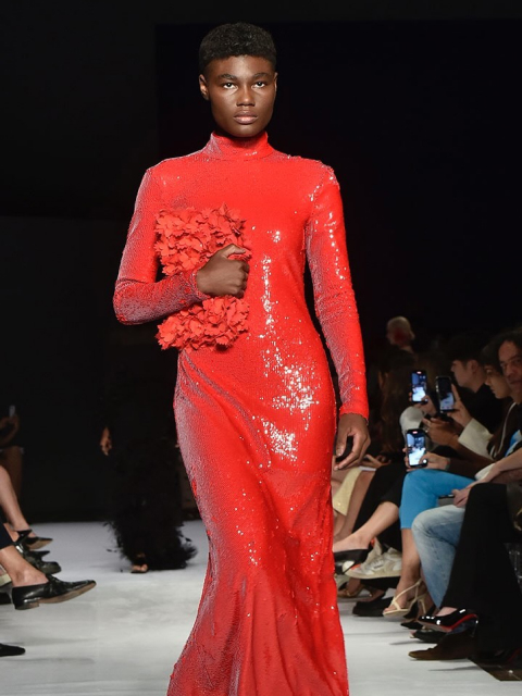 Mulher negra com longo vestido de látex vermelho segurando outra pça de roupa desfila para um público diverso,