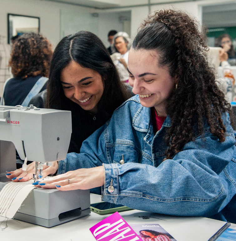 Meninas visitantes da FAAP Aberta sorrindo e praticando em uma máquina de costura